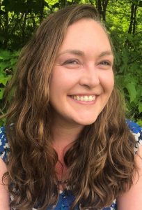 A woman with long, wavy brown hair smiles at the camera. She is wearing a blue and white patterned top and is outdoors with green foliage in the background.
