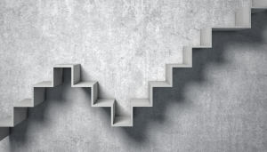 A modern, zigzag-shaped staircase is mounted on a plain concrete wall. The stairs create sharp angles and shadows, forming a geometric pattern against the gray background.