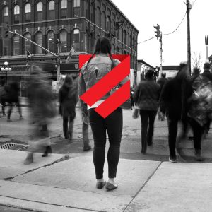 A person stands on a city sidewalk with their back to the camera while blurred pedestrians move around them. The photo is black and white. The Canadian Human Trafficking Hotline logo appears as a red, shield-shaped overlay across the person’s back, adding a branded visual element to the scene.