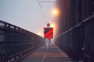 A person walks alone along a foggy bridge at dawn or dusk. Streetlights cast a warm glow through the mist, and the railings fade into the distance. The Canadian Human Trafficking Hotline logo appears as a red, shield-shaped overlay across the person’s torso.