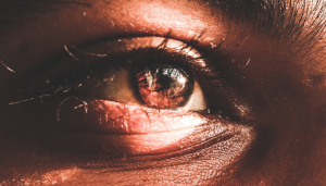 A close-up photo of a human eye, showing detailed textures of the skin, eyelashes, and iris. The lighting highlights the eyes natural colors and reflections are visible on the surface.