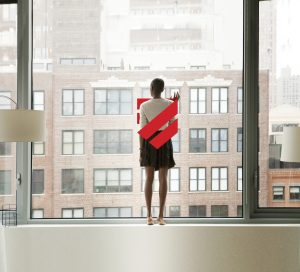 A person stands barefoot on an indoor ledge, facing a large window with their hand resting on the glass. Outside, brick apartment buildings fill the view. The Canadian Human Trafficking Hotline logo appears as a red, shield-shaped overlay across the person’s back.