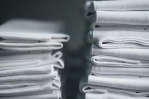 Two neat stacks of folded white fabric, such as tablecloths, placed side by side against a neutral background. The focus is on the texture and orderly arrangement of the fabric.