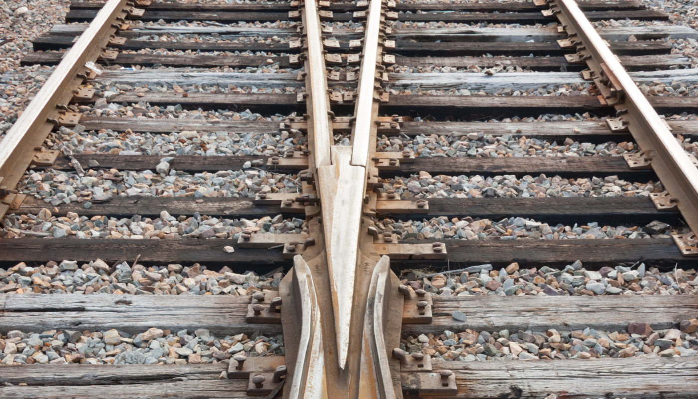 Close-up view of a railway track switch, where two sets of train tracks converge and diverge, surrounded by wooden railroad ties and gravel.