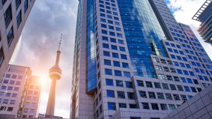 Tall modern office buildings with glass windows surround the CN Tower in Toronto, Canada, under a partly cloudy sky with sunlight shining through.