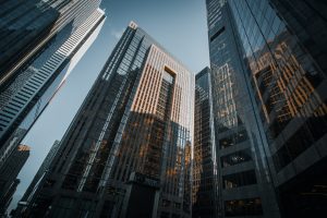 Tall glass and steel skyscrapers rise against a blue sky, their reflective surfaces capturing sunlight and surrounding buildings in an urban cityscape.