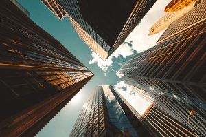 View looking up at tall glass and steel skyscrapers from ground level, with a partly cloudy blue sky above and sunlight reflecting off the buildings’ surfaces.