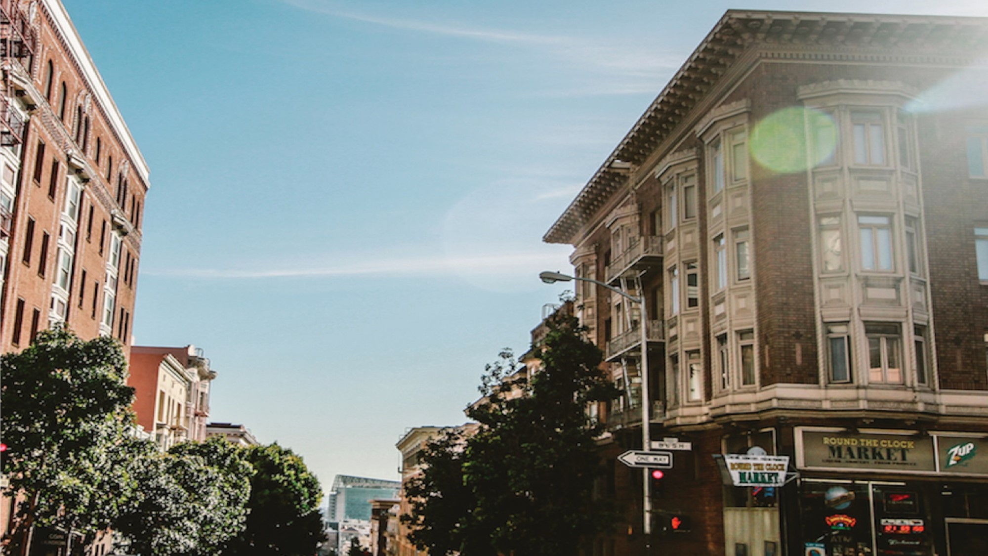 A city street scene with brick buildings, street signs, and trees under a clear blue sky. Sunlight creates lens flare on the image. A corner market is visible on the ground floor of one building.