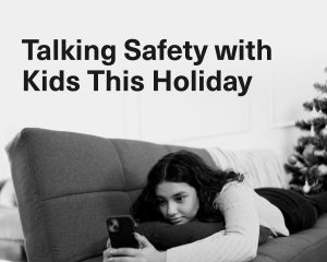 Graphic showing the text “Talking Safety with Kids this Holiday” beside a black-and-white photo of a young person lying on a couch and looking at a smartphone. A decorated holiday tree is visible in the background.