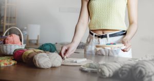 A person wearing a light green sleeveless top and white pants is holding an embroidery hoop and standing at a table with yarn, rope, and macramé supplies. Only the person’s torso and arms are visible.