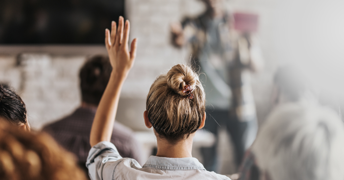 Une personne aux cheveux blonds attachés en chignon lève la main dans un groupe, face à un orateur situé à l'avant de la salle. Les autres participants sont assis et écoutent. La scène semble se dérouler à l'intérieur.