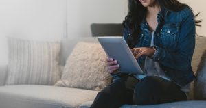A woman sits on a grey couch with textured pillows, wearing a denim jacket and dark pants, using a tablet device. The background is softly lit and slightly out of focus.
