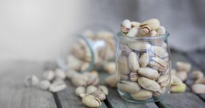 A small glass jar filled with unshelled pistachios sits on a wooden surface. Another jar lies on its side nearby, with pistachios spilling out onto the table. The background is neutral and blurred.