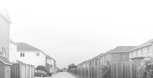 A narrow residential alley is lined with two-story houses and wooden fences on both sides. A single parked car is visible on the left. The image is in black and white with a foggy, faded sky.