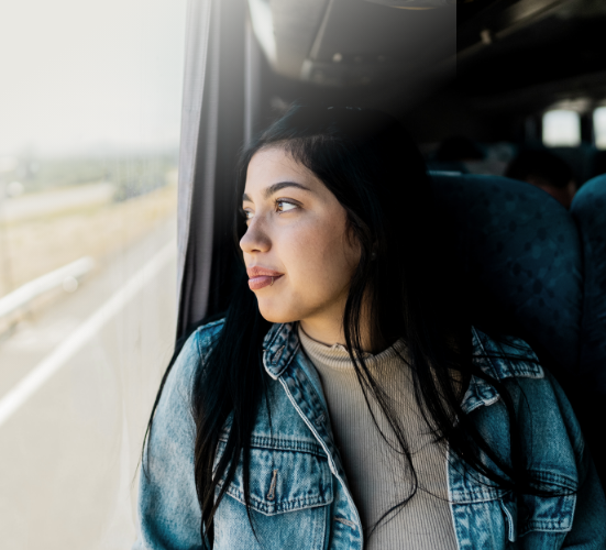 Une personne est assise dans un autobus et regarde par la fenêtre, le visage éclairé par la lumière du jour. Elle porte une veste en denim et un haut de couleur pâle.