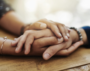 Two people sit at a wooden table, with one persons hands gently holding the others hand in a gesture of comfort or support. The background is softly blurred.