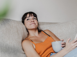 A woman in an orange tank top sits on a beige couch, holding a white mug in one hand and a smartphone in the other, looking off to the side with a slight smile.