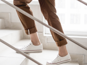 A person wearing white sneakers and brown cuffed pants is walking up a set of white tiled stairs, with a metal handrail in the foreground.