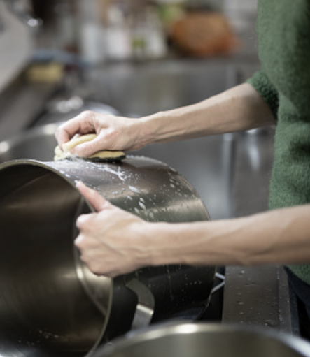 Une personne vêtue d'un chandail vert nettoie une grande casserole en métal à l'aide d'une éponge dans l'évier de la cuisine. De la mousse de savon est visible sur la casserole et l'arrière-plan est légèrement flou.