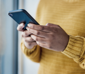 A person wearing a mustard yellow sweater is holding and using a smartphone near a window. Only the hands and part of the torso are visible.