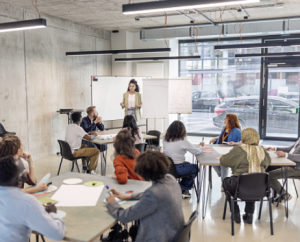 A woman stands at the front of a meeting room, speaking to a group of adults seated at tables. Participants are listening, writing, and discussing. A whiteboard and flip chart are behind her, with large windows looking out onto parked cars.