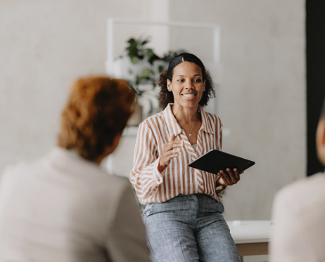 Une femme tenant une tablette est assise à un bureau et s'adresse à deux personnes assises en face d'elle dans un bureau. Elle porte une chemise rayée et un pantalon gris. Une étagère avec des plantes est visible à l'arrière-plan.