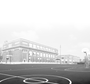A large brick school building stands behind an empty outdoor basketball court on a cloudy day. Another similar building is visible in the background, and the court has painted lines and a single basketball hoop.