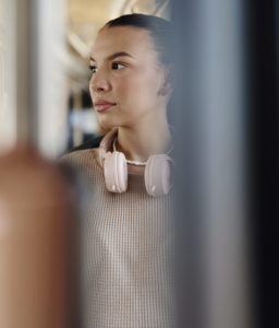 A person wearing a beige sweater with pink headphones around their neck stands on a bus, looking out the window. Out-of-focus metal poles in the foreground partly frame their face.