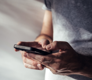 A person in a grey shirt holds a smartphone in both hands and interacts with the screen. The image is closely cropped, showing only the torso, arms, and hands.