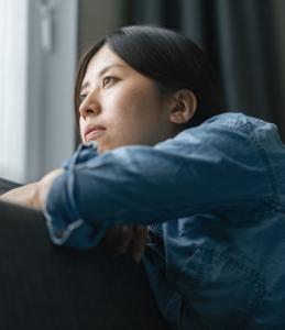 A person wearing a denim shirt sits on a couch, resting their arms on the back and looking thoughtfully out of a nearby window.
