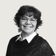 A woman with short curly hair and glasses smiles at the camera. She is wearing a collared shirt with a dark textured shawl, photographed against a light background.