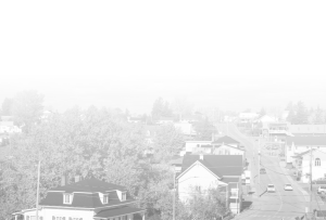 A residential street in a small town with houses, trees, and a road. The image is faded and has a white overlay, making the scene appear washed out and partially obscured.