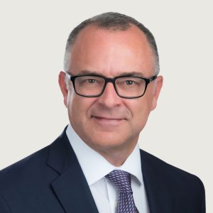A middle-aged man with short gray hair and glasses is wearing a dark suit, white shirt, and patterned tie. He is facing the camera and smiling slightly against a plain light background.