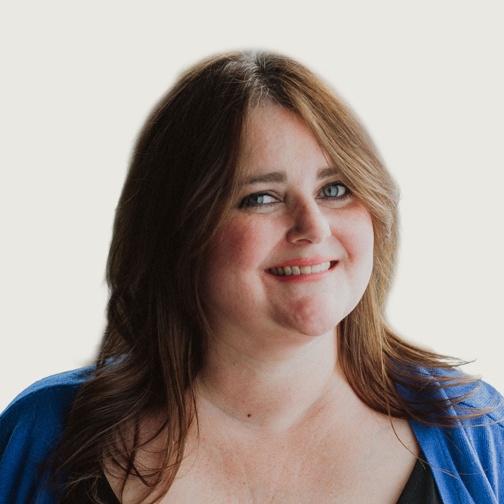 A woman with long brown hair smiles at the camera. She is wearing a blue cardigan over a black top, photographed against a light background.