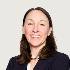 A woman with straight brown hair, wearing a dark blazer and white top, smiles while looking at the camera against a plain light background.