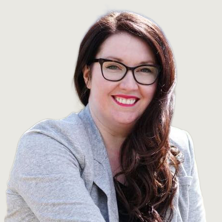 A woman with long brown hair and glasses, wearing a light gray blazer over a black top, is smiling at the camera. The background is plain and light colored.