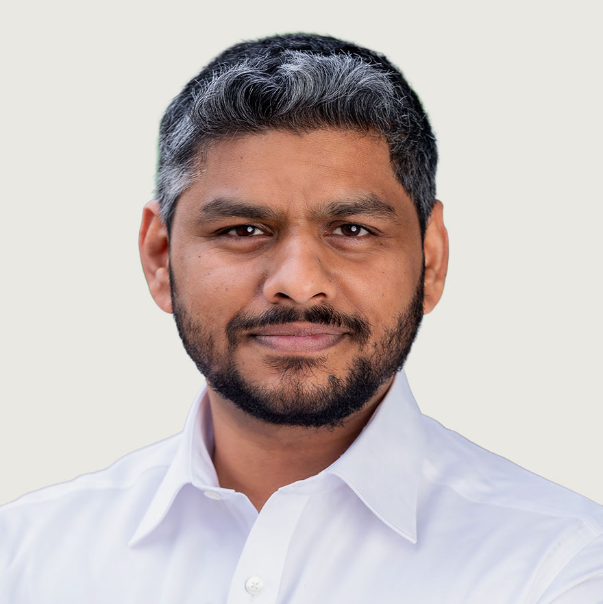 A man with short, dark hair and a beard is wearing a white button-down shirt. He is facing forward and looking at the camera with a neutral expression. The background is plain and light-colored.