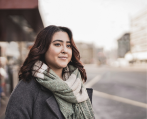 A woman with wavy brown hair wearing a gray coat and a green plaid scarf stands outdoors on a city street, looking ahead with a slight smile. The background is blurred with buildings and a road visible.