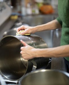 A person in a green sweater washes a large stainless steel pot with a sponge at a kitchen sink. Another pot sits nearby. The scene focuses on their hands and the cleaning process.