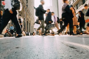 A low-angle view of a busy city street with people walking quickly in various directions. Most individuals carry bags and wear business attire, and buildings line both sides of the street. The image is slightly blurred from motion.
