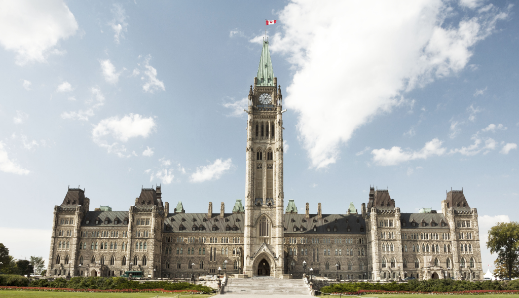 Édifice du Centre du Parlement du Canada à Ottawa, avec la Tour de la Paix au centre et un drapeau canadien flottant au sommet, sous un ciel partiellement nuageux.
