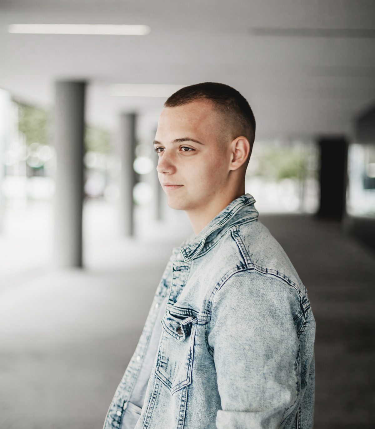 Une jeune personne aux cheveux courts, vêtue d'une veste en jean bleu clair, se tient debout dans un espace urbain extérieur, le regard tourné sur le côté. À l'arrière-plan se trouvent des colonnes en béton et une allée couverte.