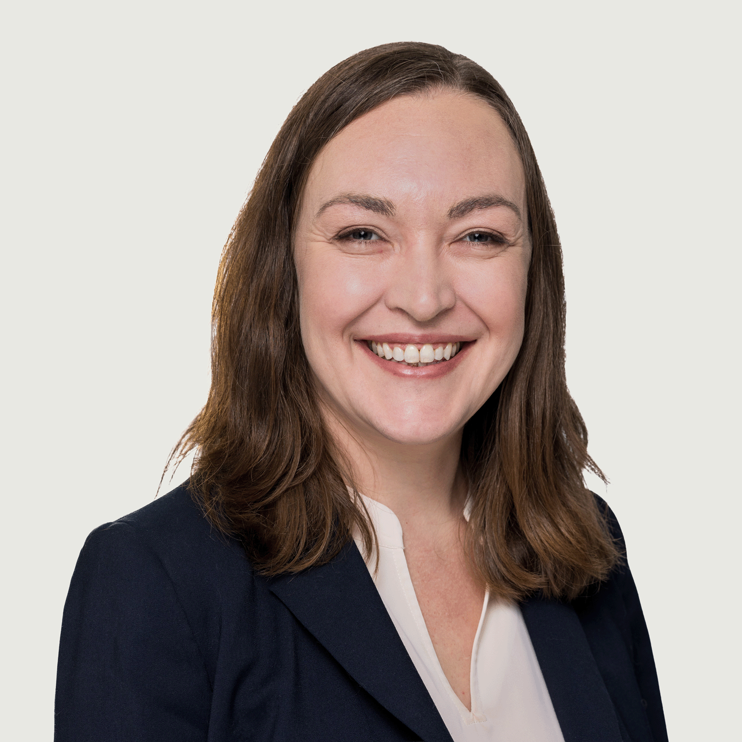 Portrait of a person with shoulder-length brown hair, smiling at the camera while wearing a dark blazer over a light blouse, against a plain light background.