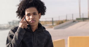 A person wearing a dark jacket stands outdoors, holding a phone to their ear and looking off into the distance.