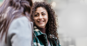 A person with curly hair and a plaid coat is smiling and looking at another person whose back is to the camera. The background is blurred.