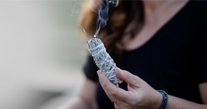 A person holding a burning sage bundle with smoke rising, wearing a black shirt and beaded bracelets. The background is blurred.