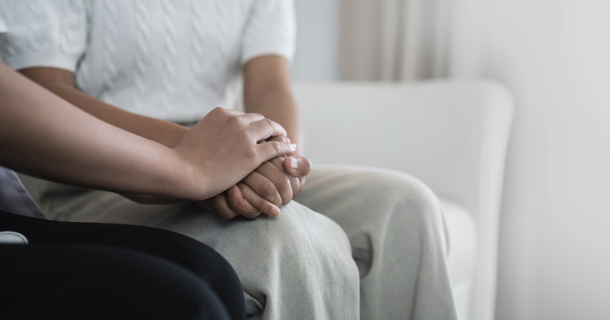 Two people sit close together indoors; one person rests their hands in their lap while the other gently places a hand on top, suggesting comfort or support. Both are wearing light-colored clothing.