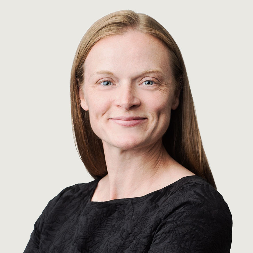 A woman with straight, light brown hair smiles slightly at the camera. She is wearing a black textured top and stands against a plain, light background.
