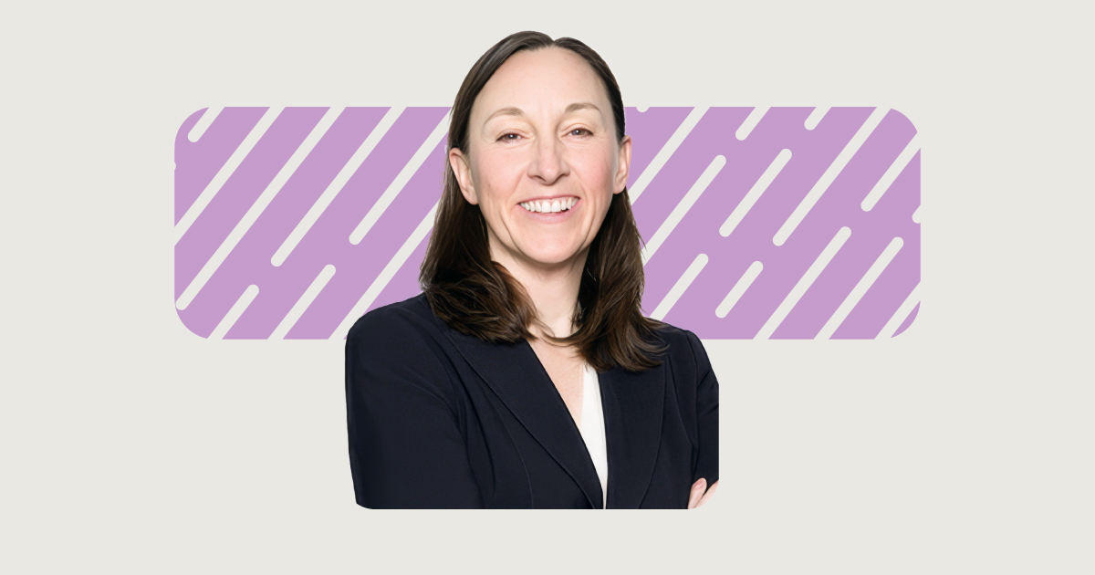 A woman with long brown hair, wearing a dark blazer and white shirt, stands smiling in front of a purple background with diagonal lines.