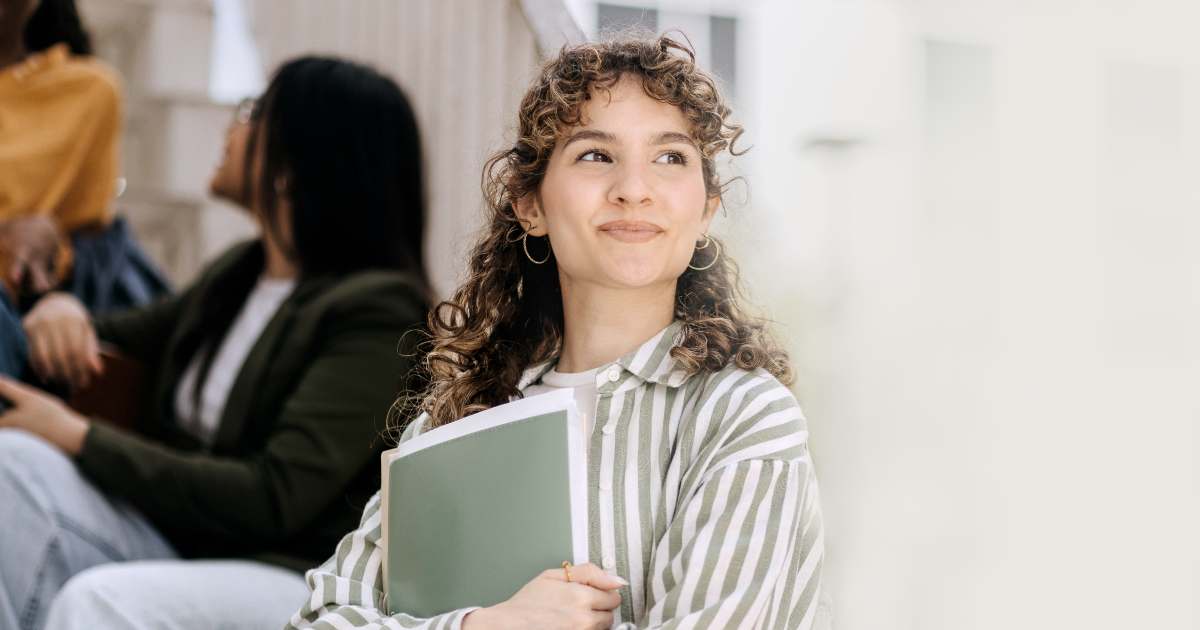 A young person with curly hair holds a folder and smiles while looking to the side. They wear a striped shirt and gold hoop earrings. Two other people sit in the blurred background, and the setting appears to be outdoors.
